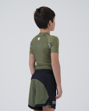 Kingz Trooper Youth Rashguard Military Green - Full Body 3/4 Back View Right Side Young Boy Athlete Short Sleeve Olive Green Crown Logo Blue Dot Sleeve Gray Side Panels Youth BJJ Kids