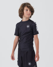 Kingz Kore V2 Youth S/S Rashguard Navy Black - Upper Body 3/4 Front View Teen Boy Athlete Short Sleeve Navy Blue White Crown Logo Circle Prominent Chest KINGZ White Text Right Sleeve Crown Logo Hem Shorts Kids BJJ Kore [color:Black]