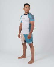 Kingz Flow Rashguard White Teal Blue - Full Body Left Side View Male Athlete Short Sleeve White Body Teal Blue Raglan Sleeves KINGZ Pink Text Chest Dark Navy Collar Matching Two-Tone Shorts Barefoot Stance BJJ Competition