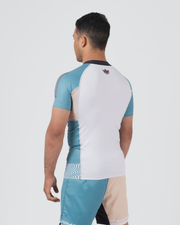 Kingz Flow Rashguard White Teal Blue - Full Body 3/4 Back View Male Athlete Short Sleeve White Back Panel Teal Blue Raglan Sleeves Dark Navy Collar Crown Logo Upper Back Matching Two-Tone Shorts Wave Pattern
