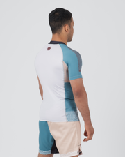 Kingz Flow Rashguard White Teal Blue - Full Body Right Side Profile View Male Athlete Short Sleeve White Body Teal Blue Raglan Sleeve Pink Crown Logo Upper Back Dark Navy Collar Matching Two-Tone Shorts Lightweight BJJ