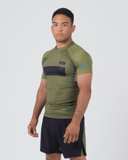 Kingz Trooper Rashguard Military Green - Full Body 3/4 Angled Front View Male Athlete Short Sleeve Olive Green Black Chest Band KINGZ Blue Text Flag Patches Gray Side Panels Black Shorts