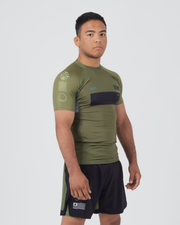 Kingz Trooper Rashguard Military Green - Full Body 3/4 Angled Front View Male Athlete Short Sleeve Olive Green Black Chest Band Blue Sunburst Dot Design Sleeve Flag Patch Black Shorts BJJ