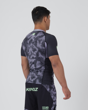 Kingz Viper Rashguard Black Purple - Full Body 3/4 Back Right Side View Male Athlete Short Sleeve Hexagonal Viper Scale Camo Pattern Green Crown Logo Black Raglan Sleeves Purple Collar Matching Shorts KINGZ Green Text