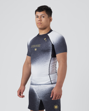 Kingz Galaxy Rash Guard Black White - Full Body 3/4 Front Left Side View Male Athlete Short Sleeve Black To White Ombre Galaxy Spatter Pattern KINGZ Gold Text Gold 4-Point Star Crown Logo Matching Galaxy Shorts
