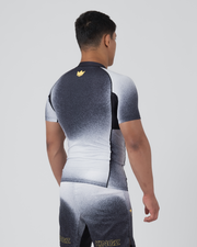 Kingz Galaxy Rash Guard Black White - Full Body 3/4 Back Left Side View Male Athlete Short Sleeve Black To White Ombre Galaxy Spatter Pattern Gold Crown Logo Upper Back Matching Galaxy Shorts KINGZ Gold Text Ankle BJJ