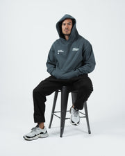 Kingz JJ Dept Hoodie - Full Body Seated Shot - Hood Up Lifestyle Look - Charcoal Blue - BJJ Streetwear Hoodie