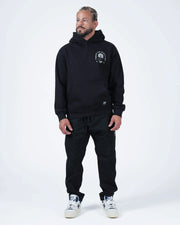 Black BJJ hoodie full length angled front shot with adult male model in all black outfit and white shoes
