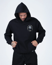 Black BJJ hoodie front lifestyle shot with adult male model wearing hood up and hands in kangaroo pocket