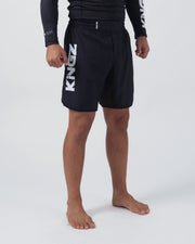 Kingz Kore V2 Shorts Black - 3/4 Angled Front View Male Athlete Black Grappling Shorts Large KINGZ White Text Left Outer Thigh Elastic Waistband Competition Cut Black KINGZ Rashguard White Text Sleeve Bare Feet Men's No-Gi BJJ MMA Kore Collection