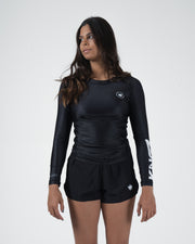 Kingz Kore V2 Long Sleeve Rashguard Black - Full Body 3/4 Front View Female Athlete Long Sleeve All Black Crown Logo Circle Chest KINGZ KIMONOS White Vertical Sleeve Text Matching Black Shorts Crown Logo Women's BJJ Kore V2