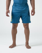 Kingz MI-K Roots Shorts Blue - Full Body Front View Male Athlete Steel Blue Teal Color KINGZ Gold Yellow Vertical Text Left Leg MI-K Circular Badge Right Thigh Competition Cut Men's Grappling Shorts No-Gi BJJ MI-K Roots Mica Galvao Limited Edition