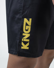 Kingz MI-K Roots Onça Pintada Shorts Black - Close Up Leg Detail KINGZ Large Bold Gold Yellow Text Left Leg Dark Charcoal Black Fabric Side Split Visible Men's Grappling Shorts No-Gi BJJ