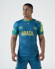 Kingz MI-K Roots Rashguard Teal Blue - Full Body Angled Front View Male Athlete Short Sleeve BRASIL Yellow Text Brazil Flag Crown Logo Green Cuffs Teal Blue Shorts Brazilian National Team