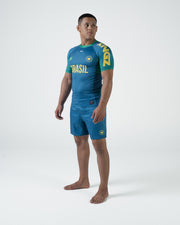 Kingz MI-K Roots Rashguard Teal Blue - Upper Body Angled Front View Male Athlete Short Sleeve BRASIL Yellow Text Crown Logo Brazil Flag Green Patterned Sleeves Collar Teal Blue Shorts
