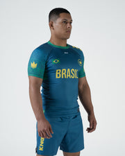 Kingz MI-K Roots Rashguard Teal Blue - Full Body 3/4 Front View Second Male Athlete Short Sleeve BRASIL Yellow Text Brazil Flag Crown Logo Green Tribal Pattern Sleeves Teal Shorts BJJ