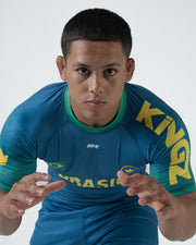 Kingz MI-K Roots Rashguard Teal Blue - Action Close Up Upper Body Male Athlete Grappling Stance BRASIL Yellow Text KINGZ Text Sleeves Green Tribal Pattern MIK Logo Dynamic Pose BJJ