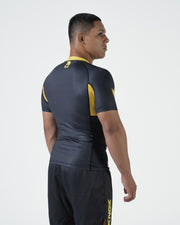 Kingz MI-K Roots Onça Pintada Rashguard Midnight Gray - Full Body 3/4 Back View Right Side Male Athlete Short Sleeve Jaguar Pattern Gold Side Stripe Gold Collar Dark Shorts KINGZ Text